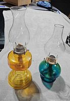 Front view of both amber and teal hurricane oil lamps with clear chimneys