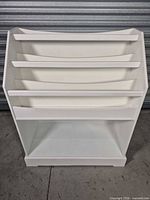 front view of white slanted display shelf showing four angled tiers and bottom cubby