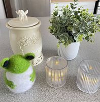 All items grouped on countertop: felt frog, cherub jar, faux eucalyptus, two lit tealights in ribbed holders