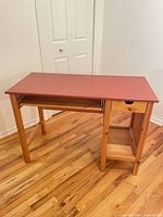 Three-quarter view of desk showing red top, keyboard tray, solid pine frame