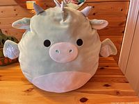 Front view of mint green Squishmallows dragon plush on wood table
