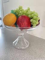 Glass pedestal compote bowl filled with assorted faux fruit (apple, lemon, green grapes) side view