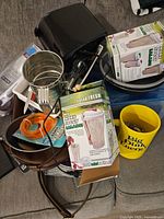 Pile of assorted kitchen items on couch including boxed Salad Fresh containers, electric grill, cup, mug, tray, flatware and cables