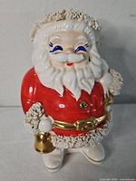 Front view of ceramic Santa coin bank showing red coat, gold bell, textured white trim