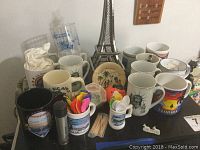 Photo showing a collection of various mugs, colorful miniature spoons, toothpicks, and the metal Eiffel Tower figurine in the background. Also visible are themed mugs with prints and a black mug holding a baseball figurine.
