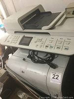Front angled view of HP color LaserJet MFP showing keypad, display screen, and paper output tray with power cord wrapped in front.