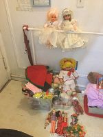 Two tall plastic dolls dressed as brides hanging on the wall, multiple small dolls and toys placed in bins and on the floor, including a Raggedy Ann doll seated in front.