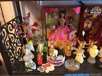 Angel figurines, Barbie doll in pink Birthday Party packaging, Star Wars Episode I collectible figure, ceramic clown bank, and decorative items on a black shelf with wrought iron rail.