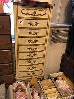 Full view of cream-colored tall lingerie chest with nine drawers, ornate brass handles, standing near other furniture and doll boxes.