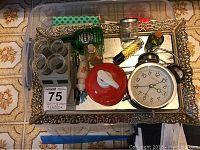 Clear overhead view of vanity items on ornate gold framed mirrored tray including alarm clock, red topped bottle, small glass jars, and small bottles of perfume and lotion