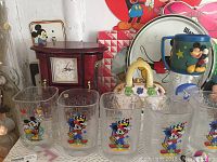 Front view of five McDonald's Millennium Edition Disney glasses with colorful Mickey Mouse designs, Disney-themed tray in background, and jewelry music box with clock