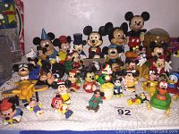 Full display of over 25 vintage Mickey Mouse and related Disney figurines arranged on a white crocheted cloth including Mickey in classic red shorts, some other characters, and small toys.