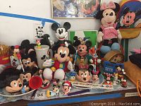 Wide view showing a collection of Mickey Mouse and Minnie Mouse items including plush dolls, figurines, mugs, and a talking phone on a tabletop.