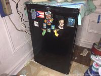 Black GE mini refrigerator with assorted magnets on the front door, placed in a corner near a wall outlet and surrounded by other household items.