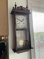Full view of wall clock hanging, showing wood case, glass door and pendulum