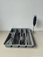 Flatware pieces sorted in grey plastic tray with black hand frother on stand
