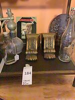 Shelf view of all seven items: pair of gold corbels, three glass bottles, metal plaque, photo frame
