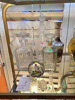 Group of five clear crystal/glass decanters and one multicolor glass paperweight on shelf