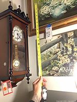 Overall view of wall clock next to two framed paintings, tape measure for scale