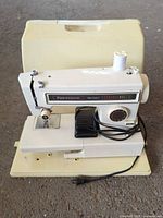 Front view of sewing machine with foot pedal, thread spool, power cord, and carrying case