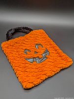 Full view of orange plush treat bag with jack-o-lantern face and black handle