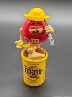 Front view of candy dispenser showing red M&M, hat, pitchfork, seagull on yellow base