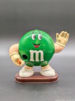 Front view of green M&M dispenser waving, showing candy opening and base