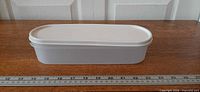 Side view of white oval Tupperware container with lid, ruler showing length