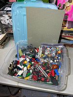 Overview of clear bin filled with mixed LEGO parts, grey baseplate leaning against lid, zipper bag of minifigures visible