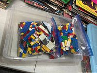 Two gallon zip bags filled with mixed LEGO pieces inside clear plastic bin