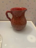 Full view of red glazed ceramic pitcher showing twisted handle and gold mottled finish