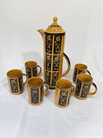 Front view of lidded coffee pot surrounded by six mugs