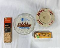 Front view showing all items: Wagnerbräu glass ashtray, Labatt's metal ashtray, Speedball Linozips box, Motomaster rubber patch kit box
