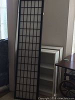 Room divider panel leaning against wall next to one bookshelf, showing size and black grid design.