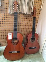 Two acoustic guitars standing vertically side by side against a netted wall, showing size difference and condition.