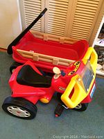 Photo showing red Fisher Price plastic wagon behind red Little Tykes tricycle indoors on carpeted floor.