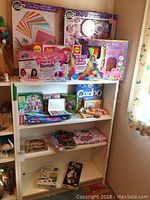 Shelving unit filled with craft kits including Knot-A-Quilt, Bath Bomb Factory, Totally Me, and Stick 'n Style along with assorted boxed games and folded fabric pieces.