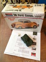 Photo showing the unopened Monogram 1936 Ford Convertible plastic model kit box and a small dark green diecast armoured car model on paper beneath the box.