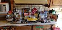 Wide view of assorted bakeware and kitchen items on countertop including metal bowls, plastic containers, boxed German clay baker, Emile Henry pot, ice trays, and vintage kitchen gadgets.
