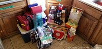 Photo showing assorted coolers, picnic baskets, thermoses, glasses, napkins on kitchen floor by wooden cabinets