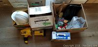 Photo showing plastic hard hats, cordless spot lights, painting kit, vent fan, and various tools in boxes on wooden floor