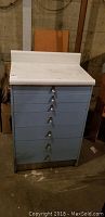 Full view of vintage medical chest showing 7 blue drawers with metal handles and light colored top surface.
