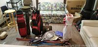 Wide shot showing two golf bags filled with clubs, tennis rackets laid in front, skis behind. Brown golf shoes visible on right.