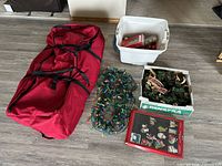 Lot overview showing red tree bag, two coils of multicolor lights, plastic tote with ornaments, wreath box and gift tag pack