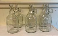Front view of six clear glass jugs showing handles and threaded mouths