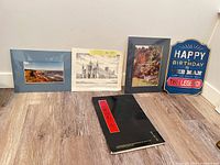 Five items leaning on wall: Cavendish photo, cathedral lithograph, village scene print, birthday sign, black portfolio