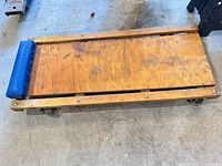 Wooden mechanic's creeper showing plywood deck, blue headrest pad, metal casters