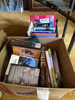 Cardboard box filled with DVDs, books, and CDs; wicker basket and additional container with more books behind