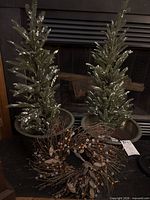 Two flocked artificial potted trees beside twig wreath with pinecones and metallic berries