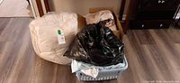 Overall view of bedspread in bag, garbage bag of linens, cardboard box and laundry basket